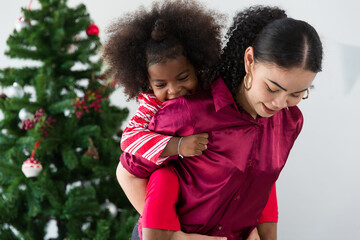 Happy African American mother piggybacking with cute curly little daughter on back. Cheerful African mom playing, having fun and spending time together over Christmas tree at home. Merry Christmas