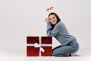Woman sitting near the big gift christmas box isolated on gray background wearing funny head hoop