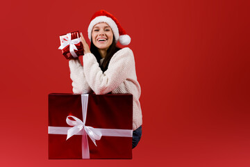 Young woman puts her ear to the christmas present wrapped in red paper sitting near big gift box, isolated on red background wearing red sanat hat