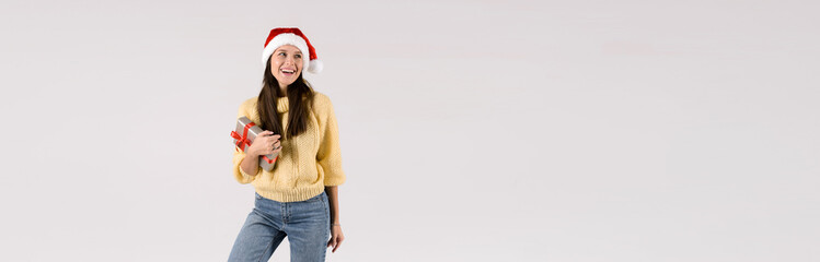 Excited young woman in red santa hat holding gift box isolated on the gray background. Banner size, empty space