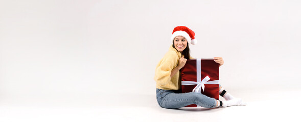 Young beautiful woman in red santa hat and yellow sweater sitting near big christmas gift box and looking to the side isolated on white background. Banner size, empty space