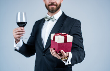 Elegant man with unshaven face cropped view in holiday festive attire hold red wine glass and gift box grey background, celebration