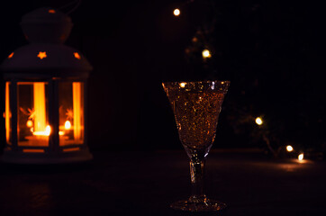 Fototapeta na wymiar Glass with white wine with garland lights and candle lantern on the background. dark picture. Burning candle. Golden beverage in the glass. Beautiful lights reflected in the glass.