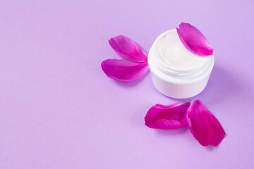 Jar with white soft cream with peony oil essential and floral petals in the corner of pink background