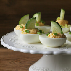 Spicy deviled eggs garnished with cucumber and leek on white plate. Wooden rustic background