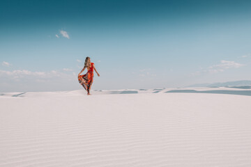 Naklejka na ściany i meble A girl in a long flowing red skirt posing in the white gypsum sand dunes of White Sands National Park, New Mexico, USA 