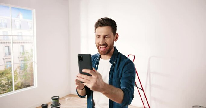 Portrait of Caucasian cheerful young bearded man in empty apartment repairing room videochatting online on smartphone in good mood. Renovation and improvement concept. Redesigning house concept