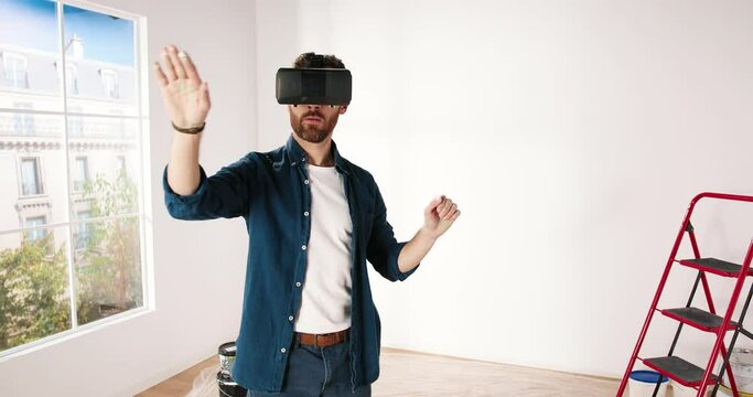 Camera approaching young Caucasian guy wearing VR glasses, scrolling with hands in air using futuristic innovative technology choosing home design. Repair and renovation concept. Virtual reality