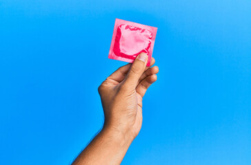 Young hispanic hand holding condom over isolated blue background.
