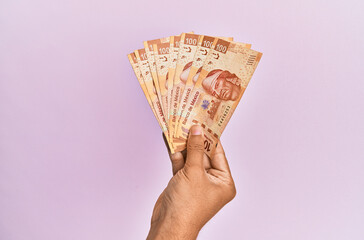Hispanic hand holding 100 mexican pesos  banknotes over isolated pink background.