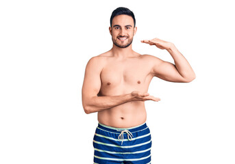 Young handsome man wearing swimwear gesturing with hands showing big and large size sign, measure symbol. smiling looking at the camera. measuring concept.