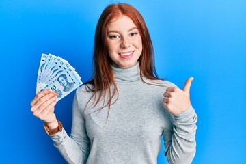 Obraz premium Young red head girl holding 10 yuan chinese banknotes smiling happy and positive, thumb up doing excellent and approval sign