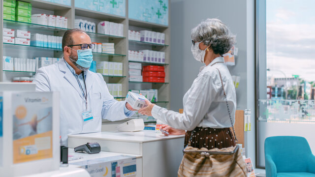 Pharmacy Drugstore Checkout Cashier Counter: Latin Pharmacist and Senior Woman Using Contactless Payment Credit Card to Buy Prescription Medicine, Vitamins. People Wearing Protective Face Masks.