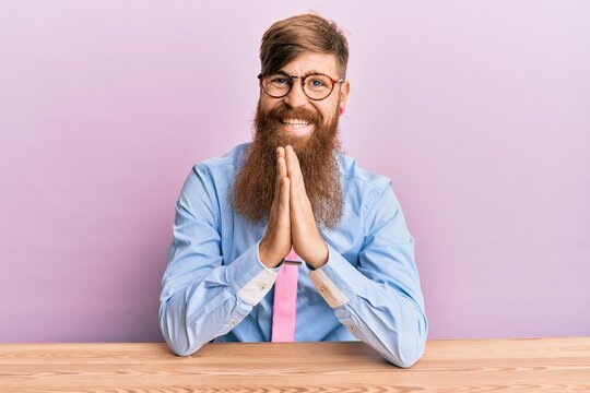 Young irish redhead man wearing business shirt and tie sitting on the table praying with hands together asking for forgiveness smiling confident.