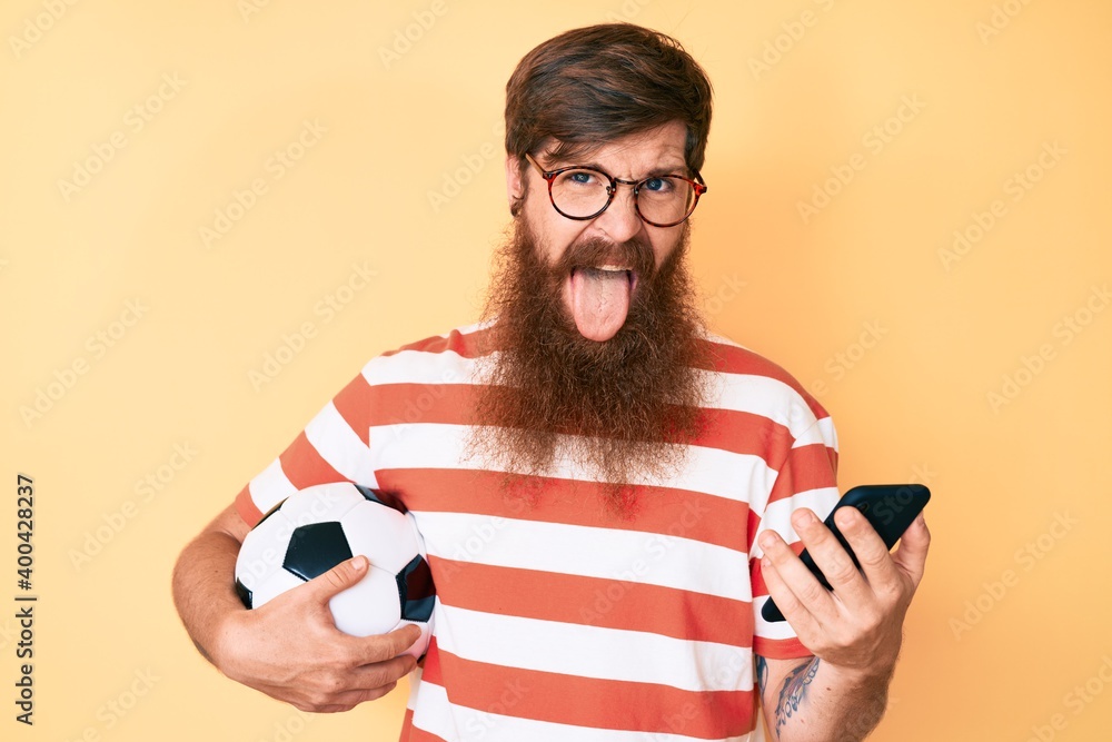 Poster handsome young red head man with long beard holding football ball looking at smartphone sticking ton