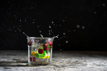 Fresh berries splash in glass of water with a spray of water droplets in motion, mint, lemon. Isolated on grey textured table with black background. Selective focus, copy space.