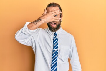 Handsome man with beard and long hair wearing business clothes peeking in shock covering face and eyes with hand, looking through fingers with embarrassed expression.