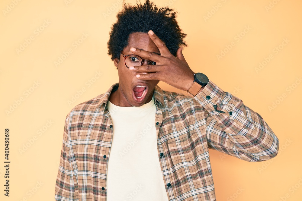 Poster Handsome african american man with afro hair wearing casual clothes and glasses peeking in shock covering face and eyes with hand, looking through fingers afraid