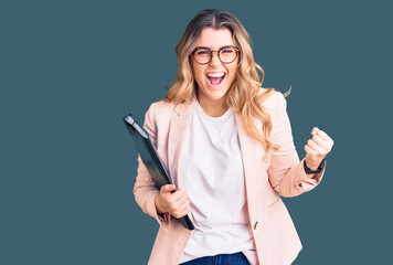 Young caucasian woman wearing business clothes and glasses holding binder screaming proud,...