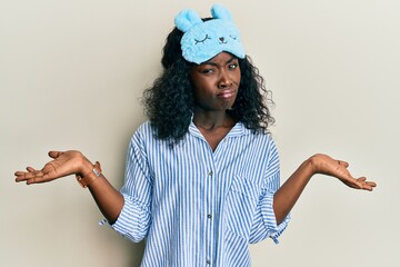 Beautiful african young woman wearing sleep mask and pajama clueless and confused expression with...
