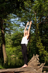 Vertical view of a young woman practicing yoga in a beautiful park outdoors. She is standing stretched arms upward on a large fallen tree practicing Virabhadrasana, or Warrior Pose