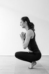 Fototapeta premium Side view of the tiptoe asana or yoga pose, with hands together at center of chest. A brunette woman in her 30’s practicing yoga at home.