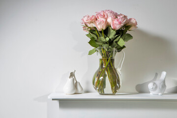 Bouquet of pink roses in a glass figured vase on a white fireplace console. A faience figurine of two pears and a bird as an interior decoration.