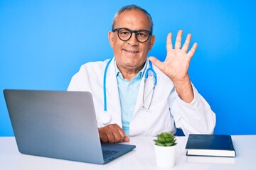 Senior handsome man with gray hair wearing doctor uniform working using computer laptop showing and pointing up with fingers number five while smiling confident and happy.