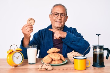Senior handsome man with gray hair sitting on the table eating breakfast in the morning smiling happy pointing with hand and finger
