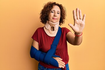 Beautiful middle age mature woman wearing cervical collar and arm on sling with open hand doing stop sign with serious and confident expression, defense gesture