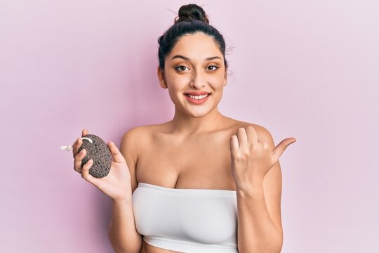 Beautiful middle eastern woman holding pumice stone pointing thumb up to the side smiling happy with open mouth