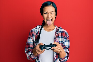 Beautiful hispanic woman playing video game holding controller sticking tongue out happy with funny...