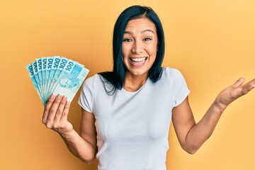 Beautiful hispanic woman holding 100 brazilian real banknotes celebrating achievement with happy...
