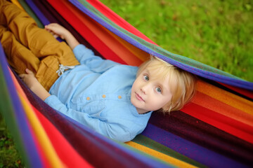 Fototapeta na wymiar Cute little blond caucasian boy having fun with multicolored hammock in backyard or outdoor playground. Summer active leisure for kids. Child swinging and relaxing in hammock.