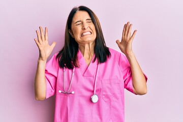 Middle age brunette woman wearing doctor uniform and stethoscope celebrating mad and crazy for success with arms raised and closed eyes screaming excited. winner concept