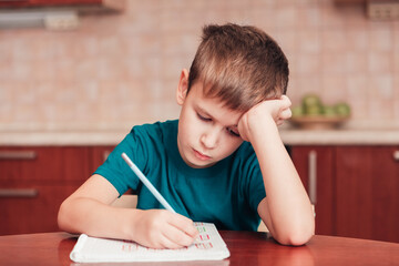 Young schoolboy learn hard at homework sitting by table in kitchen with his head on his hand and writing lessons in notebook