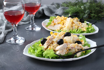 Festive salad with pineapple, baked chicken, corn and black olives on white plates and two glass of red wine on grey background. Horizontal format
