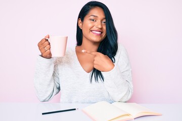 Beautiful latin young woman with long hair reading a book and drinking a coffee smiling happy...