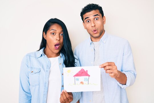 Beautiful latin young couple showing house proyect on a paper scared and amazed with open mouth for surprise, disbelief face