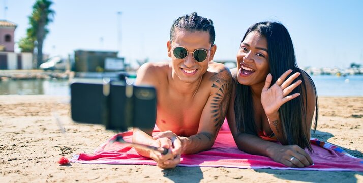 Young latin couple making selfie by the smartphone lying on the sand at the beach.