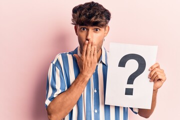 Young hispanic man holding question mark covering mouth with hand, shocked and afraid for mistake....