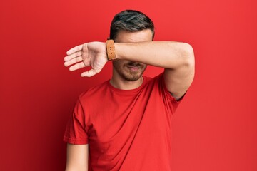 Handsome caucasian man wearing casual red tshirt covering eyes with arm, looking serious and sad. sightless, hiding and rejection concept