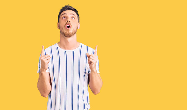 Handsome young man with bear wearing casual clothes amazed and surprised looking up and pointing with fingers and raised arms.