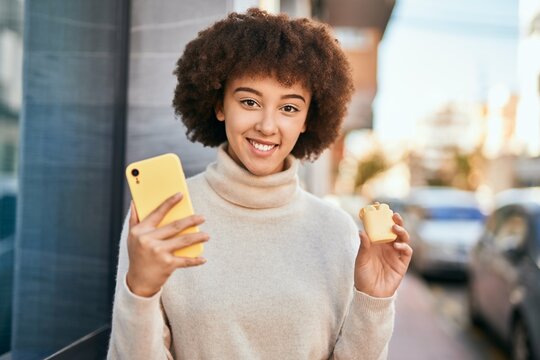 Young hispanic girl smiling happy using smartphone and holding earphones at the city.