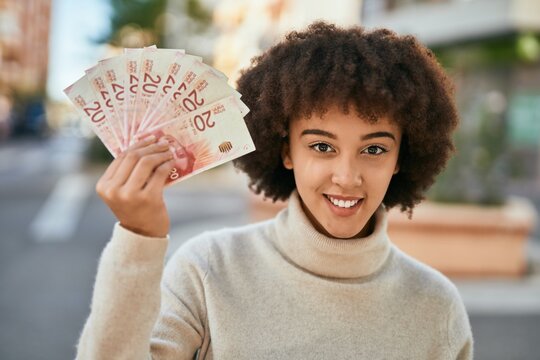 Young hispanic girl smiling happy holding israel shekels at the city.