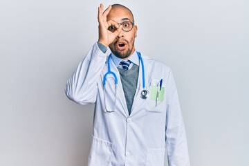 Hispanic adult man wearing doctor uniform and stethoscope doing ok gesture shocked with surprised face, eye looking through fingers. unbelieving expression.