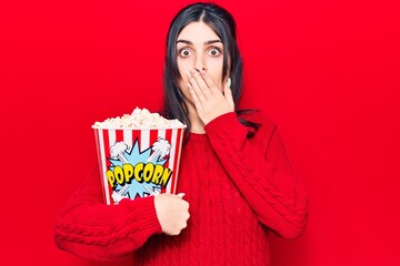 Young beautiful girl eating popcorn covering mouth with hand, shocked and afraid for mistake....