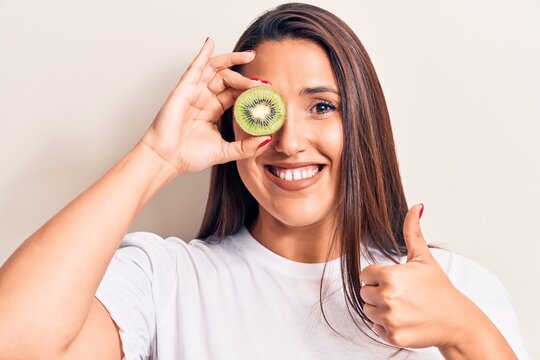 Young beautiful brunette woman holding kiwi smiling happy and positive, thumb up doing excellent and approval sign