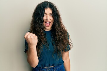 Young brunette woman with curly hair wearing casual clothes angry and mad raising fist frustrated and furious while shouting with anger. rage and aggressive concept.