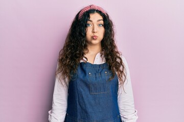 Young brunette woman with curly hair wearing cleaner apron and gloves puffing cheeks with funny face. mouth inflated with air, crazy expression.